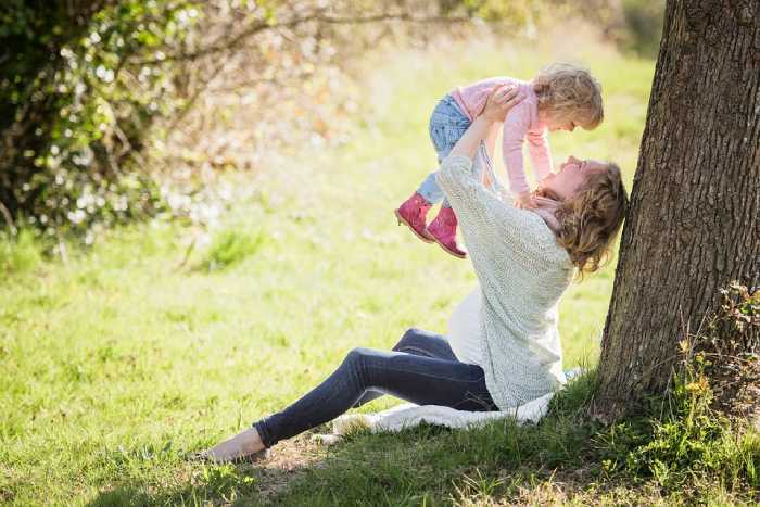 mother holding her baby above her hand and laughing while sitting against a tree